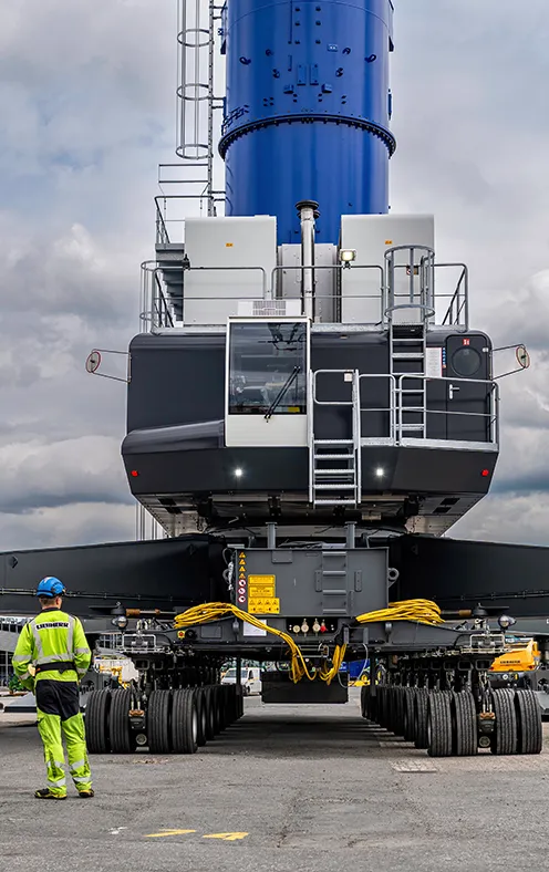 Luftaufnahme eines großen Schwerlastfahrzeugs mit vielen Rädern, das eine hohe blaue Industrieanlage transportiert; ein Arbeiter in Warnkleidung steht daneben 