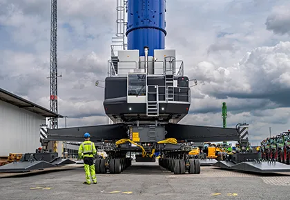 Aerial view of a large heavy transport vehicle with many wheels carrying a tall blue industrial structure; a worker in safety gear stands beside it 